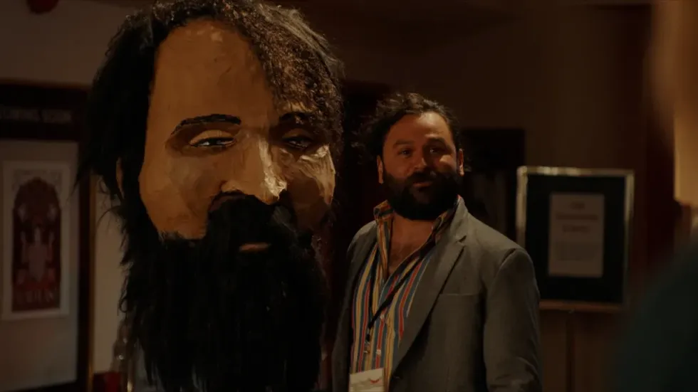 A man standing next to a paper mache head of himself, 'Hey Viktor!'