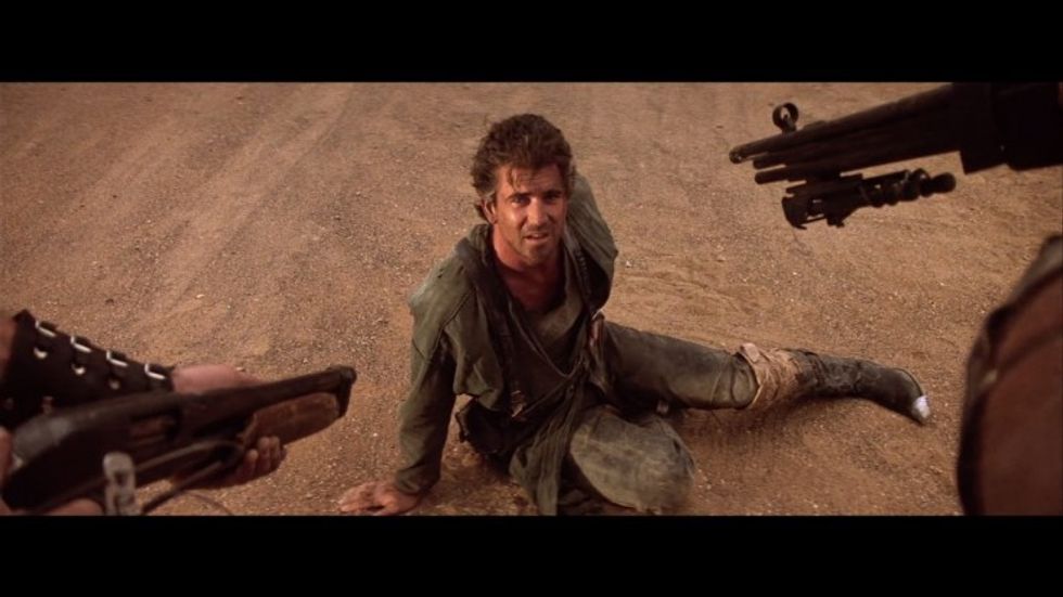 Max, played by Mel Gibson, on the ground while two guns are pointed at him in 'Mad Max'
