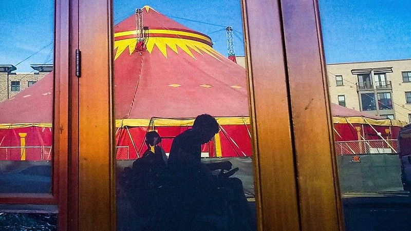 In a reflection of an unmarked storefront is a grayish silhouette of a man using an electric wheelchair. Behind the man is a spectacular red and yellow circus tent.
